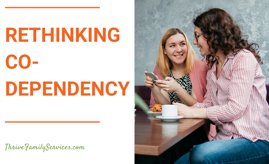 Orange text on a white background that reads "Rethinking Codependency" to the left of a photo of two women sitting at a table in front of a phone and laptop, looking at one another. | Centennial Colorado Couples Counseling