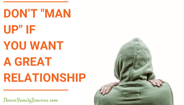 Orange text that reads "Don’t Man Up If You Want a Great Relationship" to the left of a photo of a person wearing a green hooded sweatshirt from behind. their arms are wrapped around themself and you can see their hands on their shoulders. the background, which they are facing, is white. | Englewood CO Couples Therapy