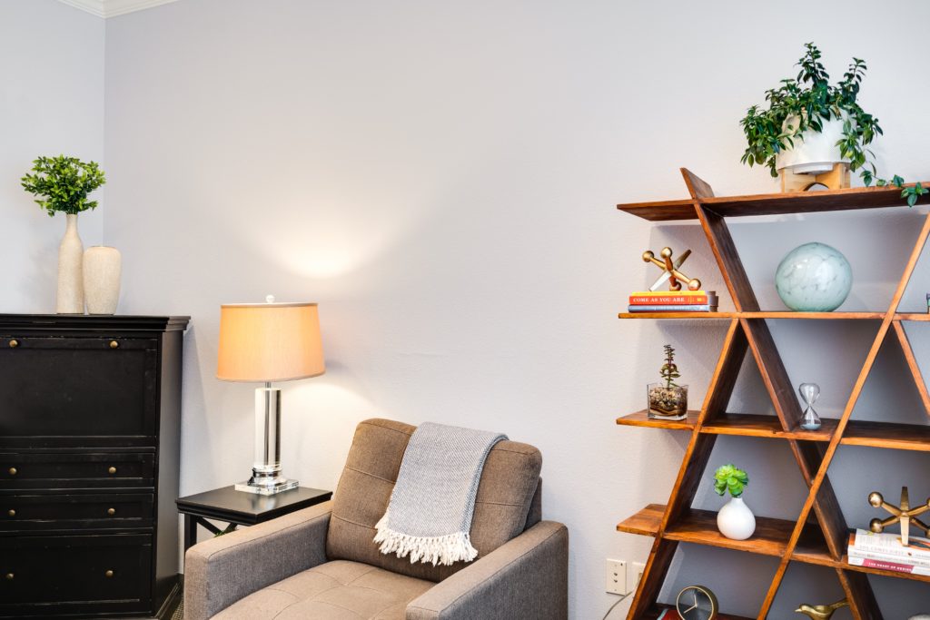 One of the therapy rooms at Thrive Family Services. We can see a white wall, against which are a geometric wooden bookcase, a gray armchair, a small black table with a lamp, and a tall black dresser. | Englewood Colorado Therapy for Adults