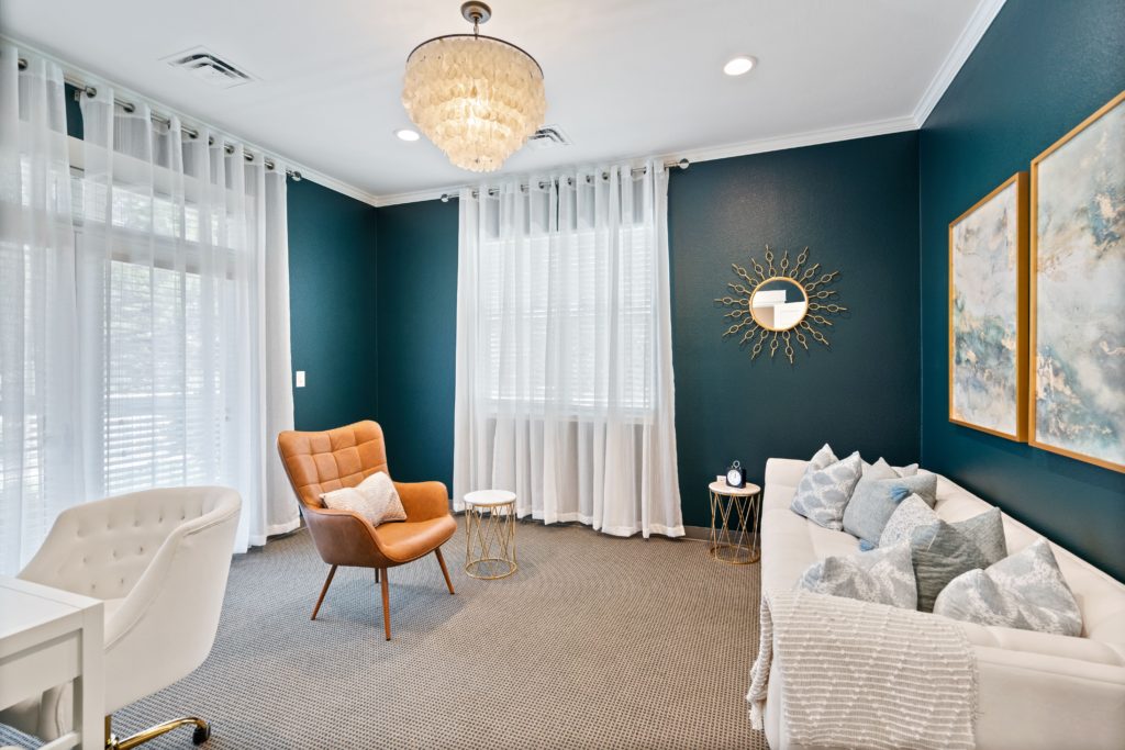 A photo of one of the therapy rooms at Thrive Family Services. The walls are a dark teal blue, and there are white curtains and a tan rug. There is a white couch with pillows, a rust colored chair, and a white desk and chair in the corner. | Centennial Colorado Couples Therapy