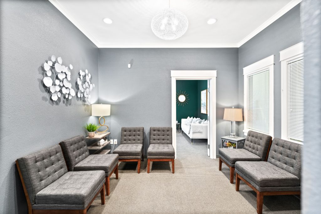 A photo of the waiting room at Thrive Family Services. The walls are gray and the rug is beige. The chairs are dark gray, sitting along the edge of the room on 3 sides. In the back we can see inside one of the therapy rooms. | Centennial Colorado Family Therapy