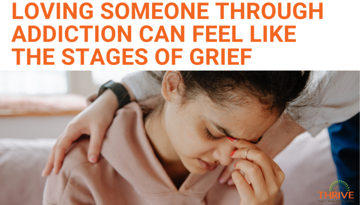 A graphic that reads "Loving Someone Through Addiction Can Feel Like the Stages of Grief" In dark orange text on a white background, above a stock photo of a non-white woman in a pink hoodie who is holding the bridge of her nose like she's in distress. Behind her someone has their arms on her shoulders in comfort. | Addiction Counseling in Centennial Colorado